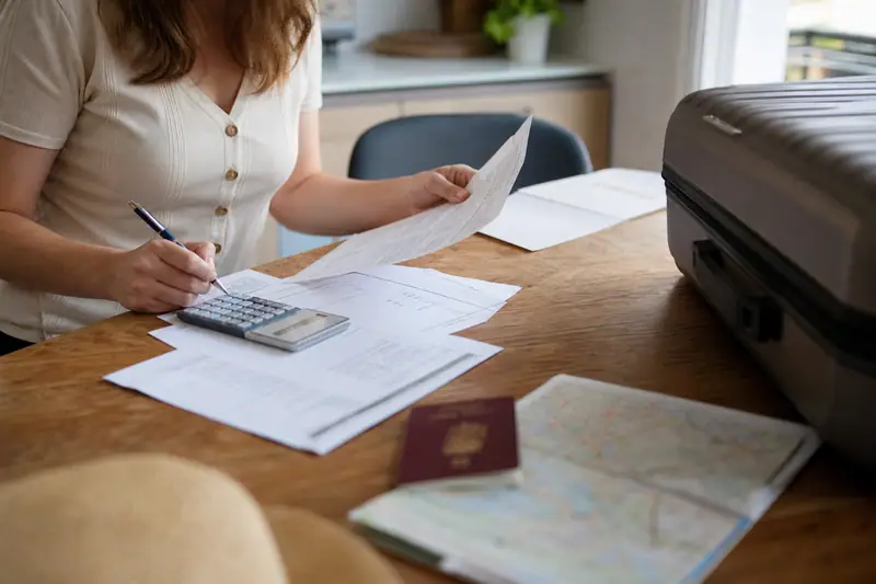 Une femme remplit des documents à table, avec passeport, carte et valise noire ouverte