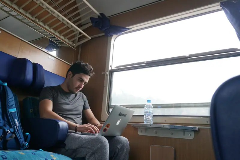Un homme assis dans un train travaille sur un ordinateur près d’un sac à dos bleu
