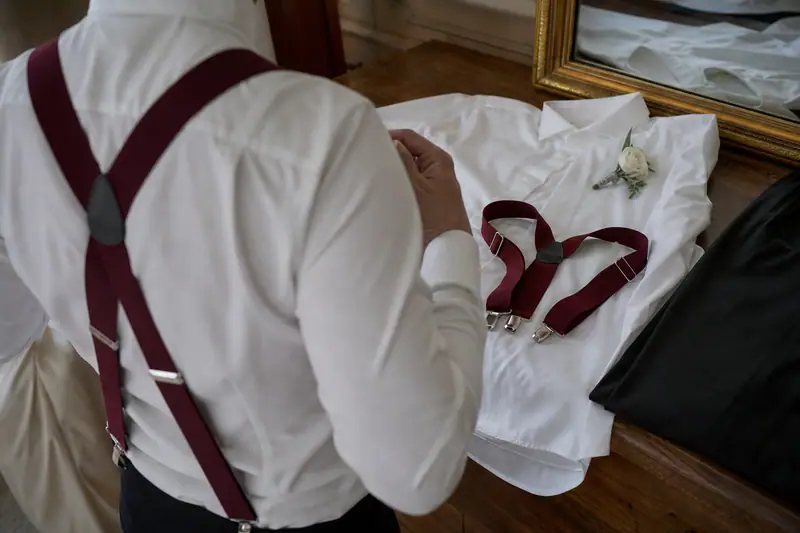 Une personne en chemise blanche porte des bretelles bordeaux devant une tenue posée sur un lit