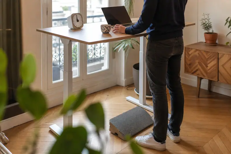 Une personne travaille debout sur un bureau blanc avec ordinateur portable près d’une fenêtre