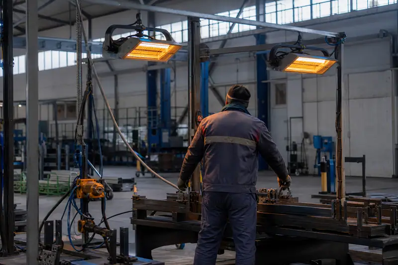 Un ouvrier manipule une machine métallique sous deux panneaux chauffants orange suspendus dans un atelier industriel