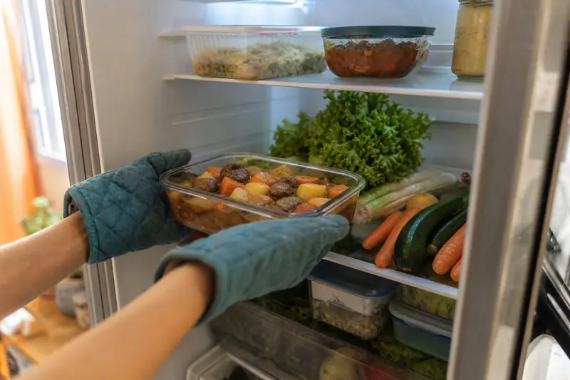 Des mains gantées rangent un plat en verre rempli de légumes dans un réfrigérateur ouvert