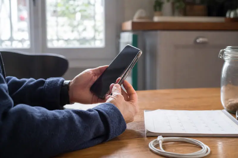 Une personne tient un smartphone noir au-dessus d'une table en bois, près d'un câble blanc