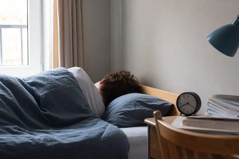 Adolescent allongé sous une couette bleue, tête sur un oreiller près d'un réveil rond
