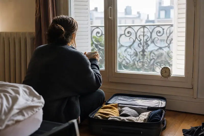 Une femme assise près d'une fenêtre tient une tasse devant une valise ouverte au sol
