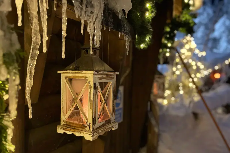 Lanterne vitrée suspendue sous des stalactites, près d’une façade enneigée éclairée de guirlandes
