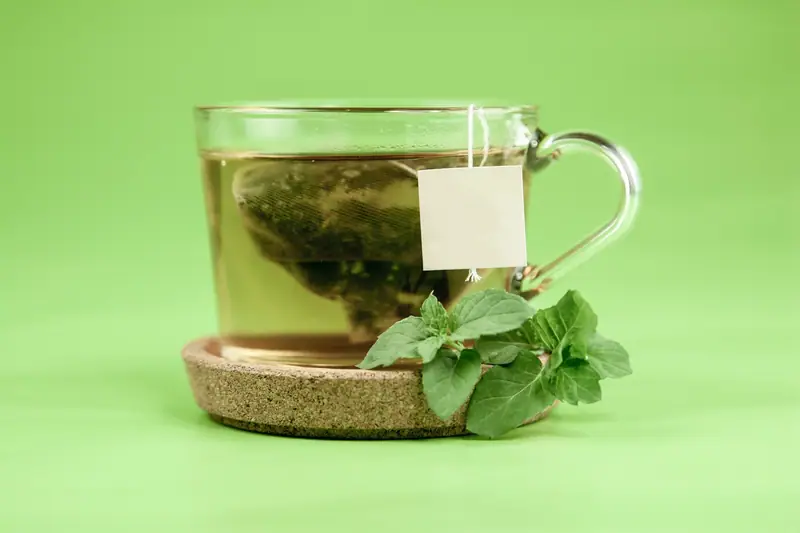 Tasse transparente de thé vert avec sachet, posée sur une soucoupe près de feuilles de menthe