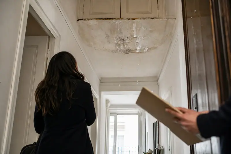 Une femme observe un plafond taché et écaillé dans un couloir, tandis qu’une main tient un carton