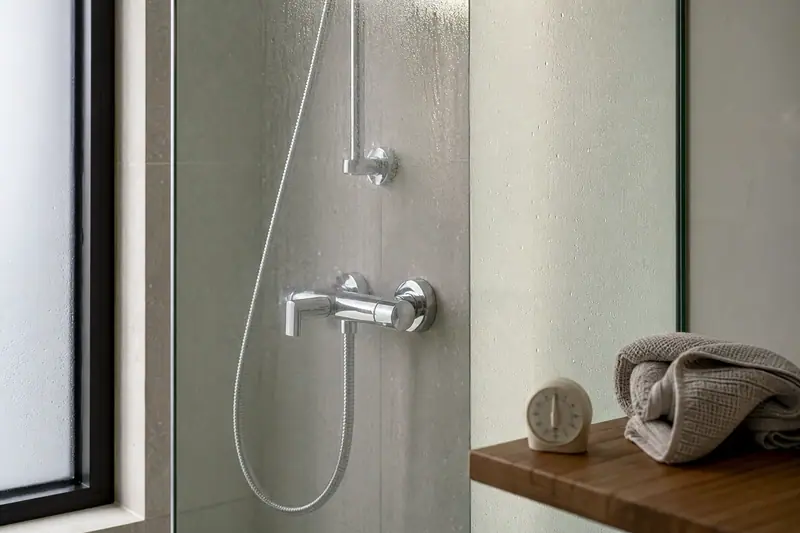 Douche vitrée avec robinetterie chromée, serviette grise roulée et réveil sur une tablette en bois