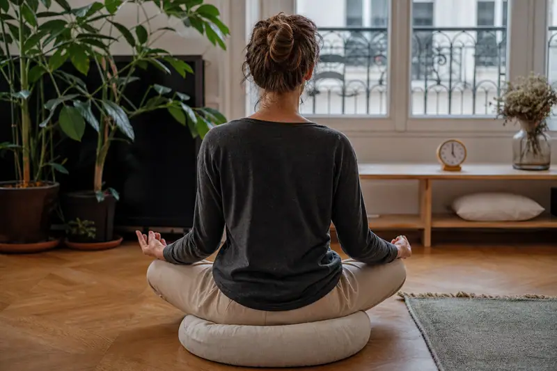 Femme assise en tailleur sur un coussin, face à une fenêtre dans un salon