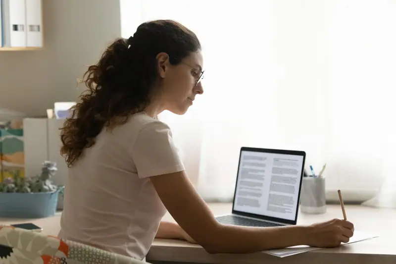 Femme assise à un bureau, écrivant sur papier devant un ordinateur portable affichant du texte