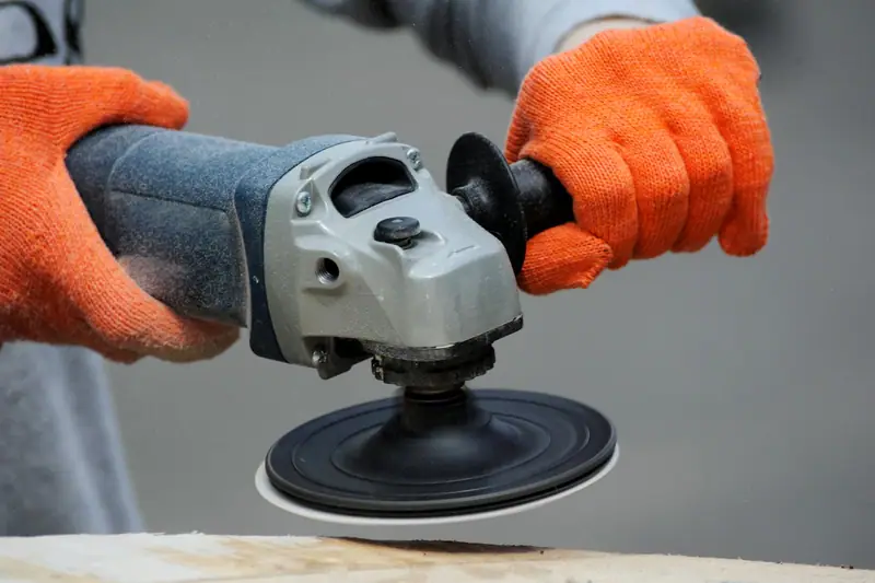 Deux mains gantées d’orange tiennent une ponceuse électrique au-dessus d’une planche de bois