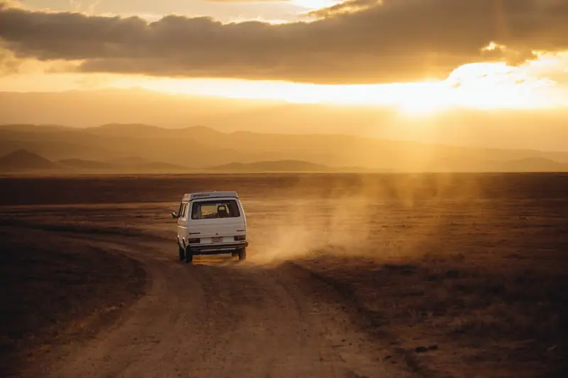 Un petit fourgon blanc roule sur une piste poussiéreuse dans un paysage montagneux au coucher du soleil