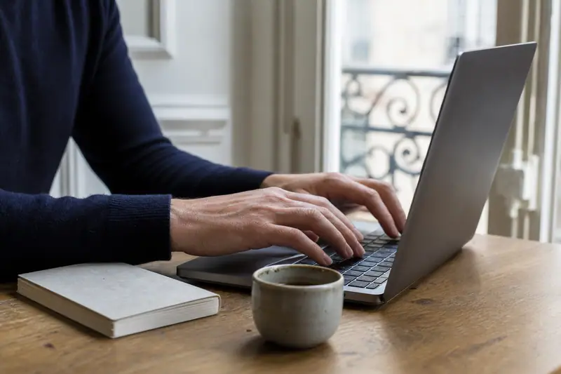 Mains d’une personne tapant sur un ordinateur portable près d’un carnet et d’une tasse
