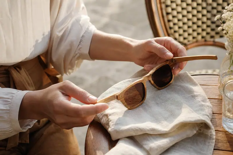 Des mains tiennent des lunettes de soleil en bois au-dessus d’un linge beige sur une table ronde