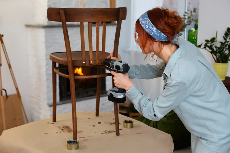 Une femme utilise une perceuse sur une chaise en bois posée sur une table protégée