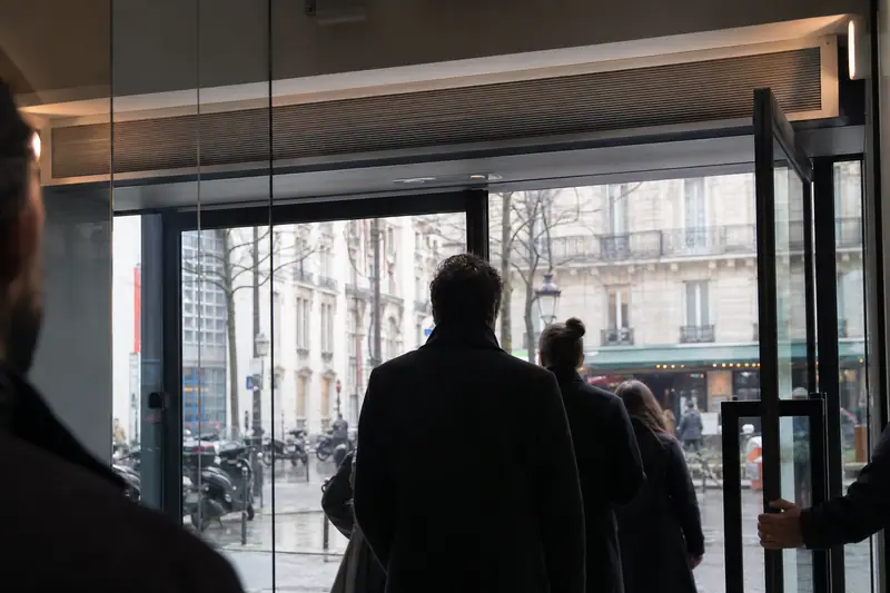 Personnes en manteaux sombres franchissant une porte vitrée donnant sur une rue pluvieuse
