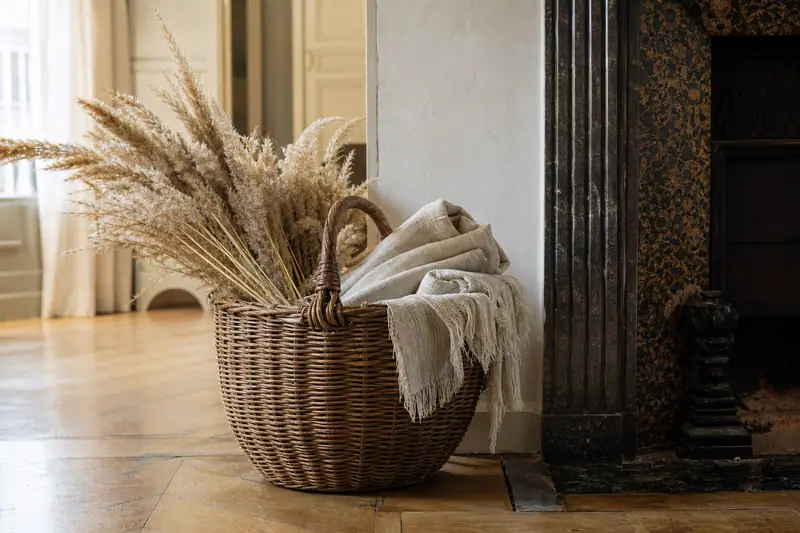 Un panier en osier contient des herbes séchées et des textiles beiges près d’une cheminée sombre