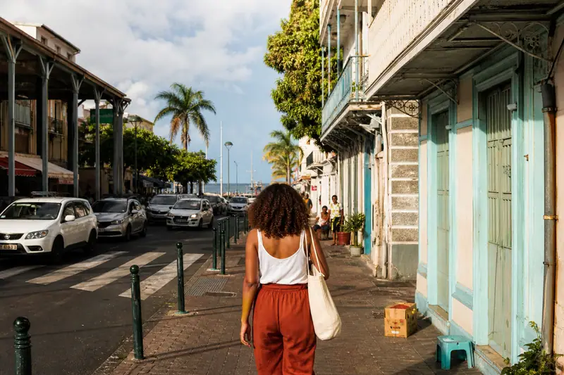 Une femme marche sur un trottoir bordé de façades turquoise et de voitures stationnées