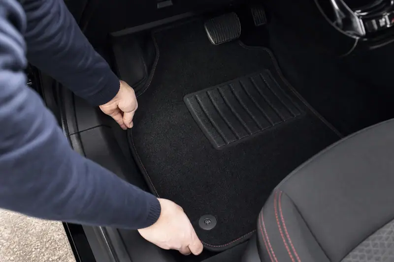 Deux mains installent un tapis noir dans le plancher d’une voiture, près d’un siège gris à surpiqûres rouges