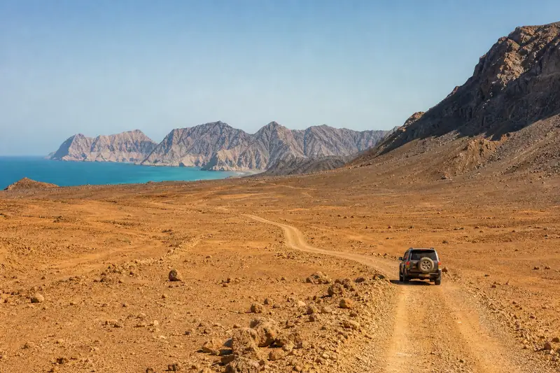 Un véhicule tout-terrain roule sur une piste de terre entre des montagnes rocheuses et une mer bleue