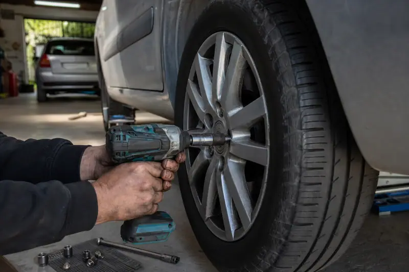Des mains utilisent une visseuse sans fil sur la roue avant d’une voiture grise dans un garage