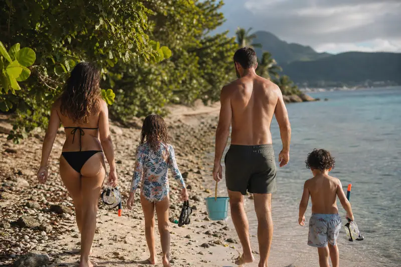 Deux adultes et deux enfants marchent pieds nus sur une plage, portant seaux et équipements de plongée