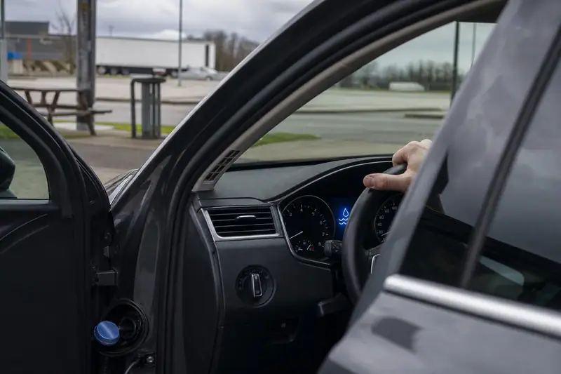 Une main tient le volant d'une voiture sombre, portière ouverte sur un parking